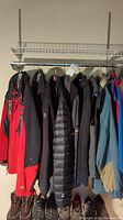 Photo showing a row of seven men’s large outerwear items hanging on a rack with footwear underneath, colors vary from red, black, blue, and grey.