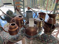 Full view of all copper kettles and watering cans arranged on glass table showing various shapes, sizes and conditions