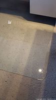Top-down photo of clear glass office chair mat on carpeted floor, showing corner and surface detail including visible dust and debris.