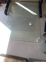 Top view of thick, heavy rectangular glass floor protector on beige carpet under office furniture, showing clear glass with rounded corners and dirt marks.