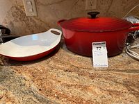 Photo of a red Le Creuset Dutch oven with lid next to a red and white baking dish on a granite countertop with a MaxSold tag in front.
