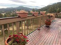 Outdoor wooden deck with two large terracotta planters and four rectangular deck planters mounted on railing, one round clay pot on deck surface, natural outdoor mountainous background