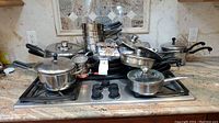 Wide view of various stainless steel pots and pans stacked on stove top with lids, showing quantity and condition requiring cleaning.