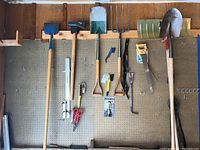 Tools hanging on a garage pegboard wall, including shovels, axe, pick, hand saws, scissors, and pruning shears with most items showing dirt and wear.