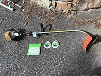 Full length image of the Stihl FS 56 trimmer laying on asphalt with user manual and two string spool packs in front.
