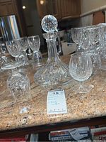 Waterford crystal decanter with stopper and 8 brandy snifters on a kitchen counter. Includes auction tag. Items are clear crystal with detailed cut patterns.