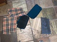 Six wool scarves arranged on tiled floor including beige plaid, solid teal, grey with stripes and fringes, cream with stripes, and dark grey scarf.