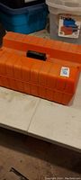 Closed orange plastic carrying case with handle and lot number tag.