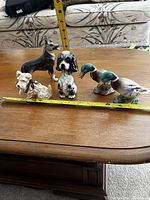 Five small animal figurines on a wooden table with measuring tape showing scale, including three dog figures and two duck figures.
