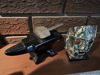 Photo showing the Avon blacksmith's anvil figurine with a small metal hammer on top alongside the abalone seashell mask on stand, placed on a wooden surface with a brick background.