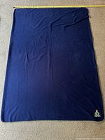 Full view of a blue fleece blanket laid flat on a carpeted floor with a measuring tape along one edge indicating approximate size.