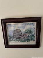 Framed watercolor painting showing the Rome Colosseum under a cloudy sky with trees and ruins nearby. Dark wood frame with textured carving.