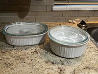 Two white Corning Ware baking dishes with glass lids on a kitchen countertop, round and rectangular shapes.