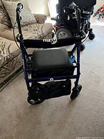 Blue Hugo Elite walker with padded seat, handle brakes, and under-seat storage pouch, shown in living room setting.