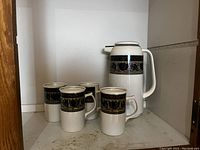 Front view of thermos carafe and four matching cups showing black and gold decorative band design.