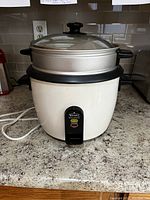 Front view of Rival rice cooker with included metal steamer basket inside and glass lid on top, placed on kitchen counter.