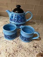 Blue floral ceramic tea set including teapot with black lid, creamer, and sugar bowl on countertop.