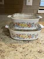 Two white CorningWare casserole dishes stacked with floral vegetable garden pattern and clear glass lids on granite counter.