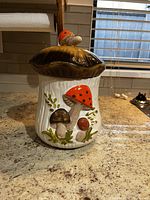 Front view of the ceramic toadstool cookie jar with brown cap lid and mushroom decorations on white body standing on countertop.
