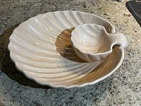 Large cream-colored ceramic shell-shaped serving dish with small attached shell-shaped dip bowl sitting on a granite countertop. Shows shape, texture, and glaze.
