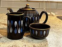 Black ceramic teapot, creamer, and sugar bowl on countertop showing teardrop pattern and gold accents