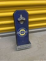 Front view of Chevrolet bottle opener with cap catcher mounted on blue board featuring Chevrolet Super Service logo and Genuine Chevrolet metal base.