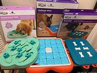Photo showing all three dog puzzle games with their original boxes in the background. The games are Puppy Hide N Slide (green), Challenge Slider (blue and orange), and Dog Casino (blue with white bone covers).