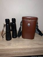 Full view of the black 8x56 binoculars alongside the brown hard case on a wooden surface showing the overall condition and contents of the lot.