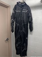 Front view of black Kamik snowmobile suit hanging on hanger in utility room