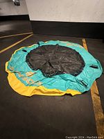 Image of the inflatable trampoline laid flat, showing the entire circular shape with its turquoise and yellow colors and central black jumping area.