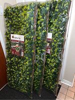Three expandable willow trellises with dense artificial laurel leaf foliage in original plastic packaging, standing upright indoors.