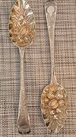 Pair of two sterling silver berry spoons displayed side by side on woven mat, showing overall design and gilded bowls