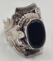 Close-up image of a chunky sterling silver ring with a polished oval black onyx stone and detailed textured silverwork featuring floral and leaf motifs around the stone.