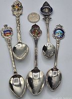 Topside view of the five souvenir spoons with colorful enamel and detailed tops showing Canadian designs and crests, placed around a Canadian quarter for scale.