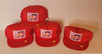 Four red mesh back trucker hats with Elvis Presley postage stamp design patches on the front, stacked in front of a plain wall
