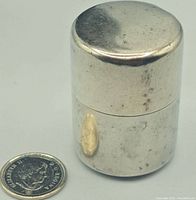 Front view of the silver canister next to a coin for size reference, showing its cylindrical shape and rounded lid.