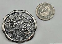 Top front view of circular sterling silver brooch with flower motif next to coin for size reference.