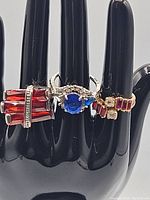 Three vintage costume rings displayed on black ring holder, showing red and blue stones