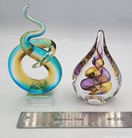 Two art glass miniatures side by side with a ruler for scale, showing a twisted loop sculpture and a teardrop-shaped glass sculpture with swirls.