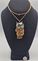 Front view of owl pendant necklace displayed on black bust, showing gold chain and green eyes on silver-tone head with gold articulated feather body.