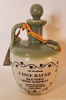 Side view of ceramic vintage Irish whiskey jug with green stopper and handle, cream body with black vintage whiskey branding and orange ribbon with tag.