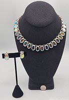 Front view of the necklace displayed on a black bust along with the earrings on a stand, showing the mirrored rainbow effect of the stones.