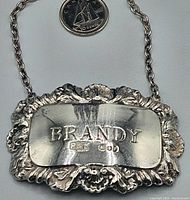 Full view of the sterling silver vintage decanter label, front face with 'BRANDY' engraving, decorative border and chain, quarter for size reference