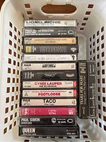 Cassette tapes in a plastic basket showing spine titles and labels for various artists including Lionel Richie, Prince and The Revolution, John Denver, George Michael, Eurythmics, Cyndi Lauper, and more.