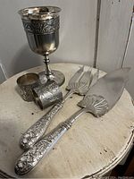 Photo of the complete lot showing a goblet, two table servers with floral handles, and two napkin rings with floral engraving.