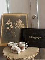 Photo showing antique sepia-tone photograph of two children sitting on a hobby horse, placed behind three German porcelain cups and a black vintage photo album labeled 'Photographs'.