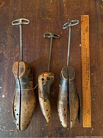 Lot contains three wooden shoe stretchers with metal crank mechanisms laid on a wood surface alongside a wooden ruler for scale, showing each stretcher’s length and condition.
