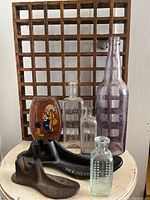 Full view of lot contents on round white table showing wooden oak floor grate, three glass bottles, vintage toffee tin, and two cast iron shoemaker forms.