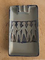 Top-down view of rectangular ceramic ashtray with relief depiction of four standing human figures and three cigarette rests along top edge. Pale blue base color with darker blue glaze accents. Shows visible glaze wear and small chips around edges.