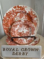 Three Royal Crown Derby Red Aves china plates displayed upright with a Royal Crown Derby display sign in front, showing the intricate red bird and floral pattern with gold trim.